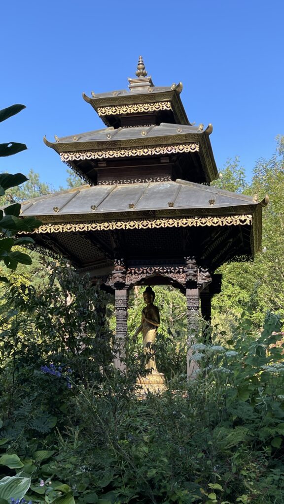 Ein mehrstufiger Pavillon mit aufwendigen Holzschnitzereien steht in einem Garten mit üppigem Grün und einem klaren blauen Himmel.