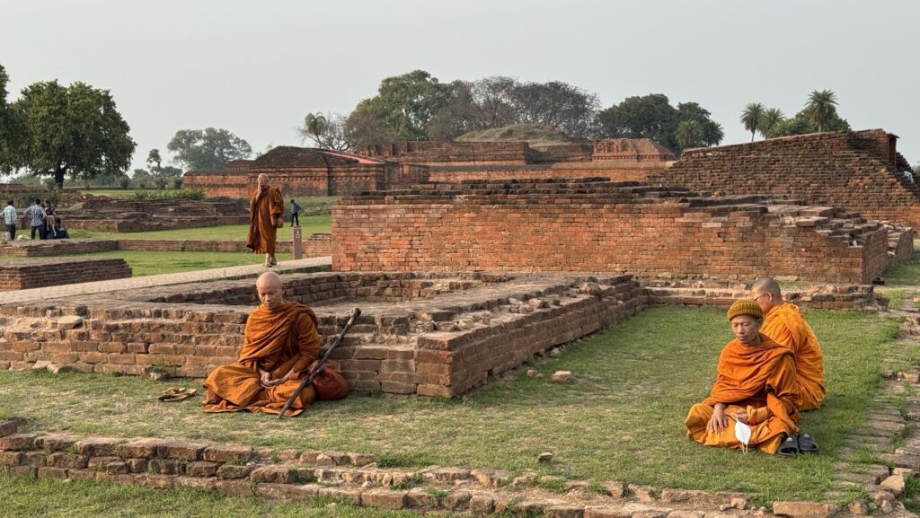 Buddhistische Pilgerreise, Nalanda. Drei Mönche in orangefarbenen Roben sitzen und meditieren zwischen alten Backsteinruinen und Grasflächen.