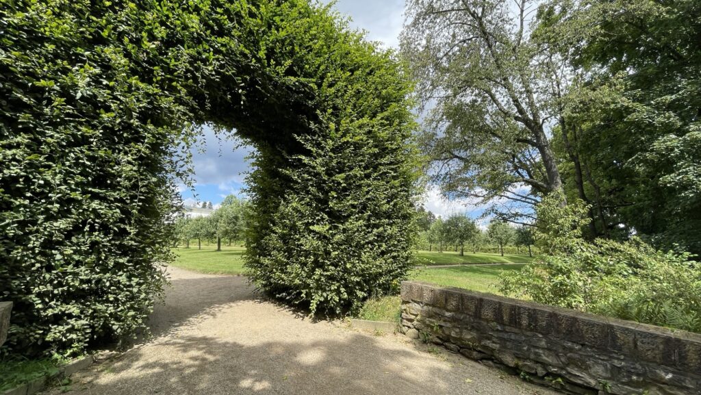 Ein Kiesweg führt durch einen grünen Torbogen aus ordentlich geschnittenen Hecken, umgeben von üppigen Bäumen und Grasflächen unter einem teilweise bewölkten Himmel.