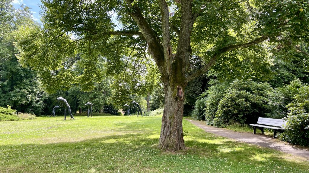 Eine Parkszene mit einem großen Baum, einer Bank und Skulpturen, die Insekten ähneln.