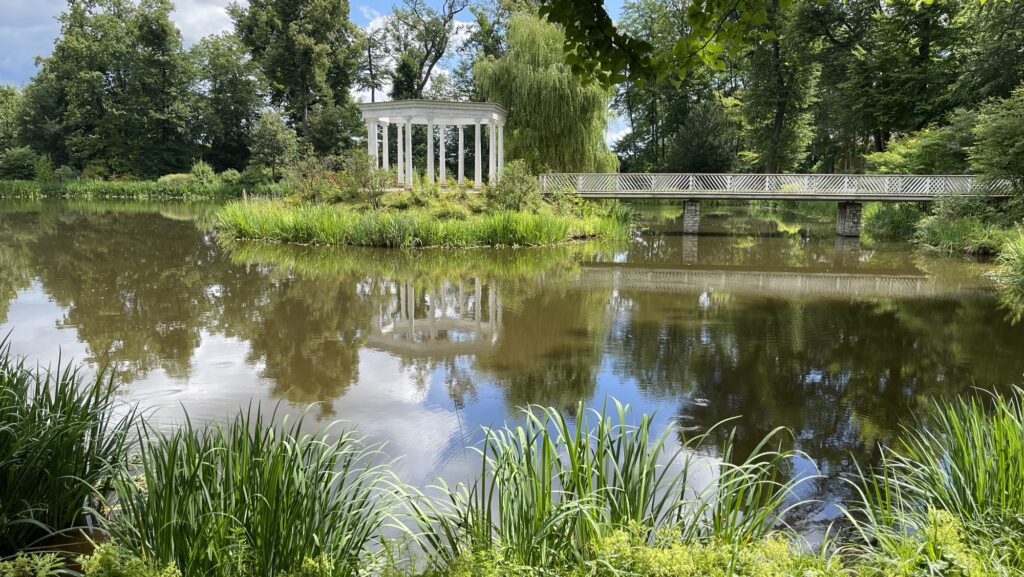 Bad Homburg, kleiner Tannenwald. Ein weißer Pavillon steht neben einem Teich mit reflektierender Oberfläche, umgeben von grünen Bäumen und Sträuchern. Unter einem teilweise bewölkten Himmel erstreckt sich eine Brücke über das Wasser.