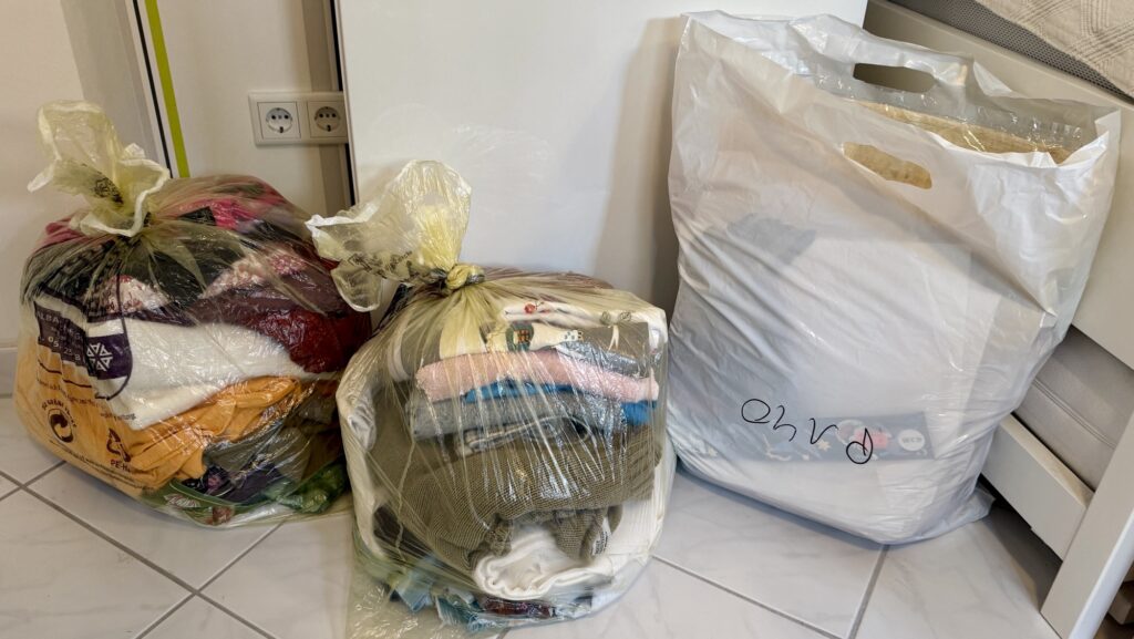 Aufräumen - Auf einem gefliesten Boden stehen drei Säcke mit Kleidung. Zwei davon sind durchsichtige Plastiksäcke, die mit verschiedenfarbiger Kleidung gefüllt sind, und einer ist ein großer weißer Sack, der mit unterschiedlichen Gegenständen gefüllt ist.