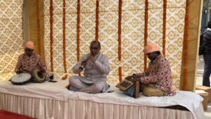 Buddhistische Pilgerreise, Safdarjung Station in New Delhi. Drei Musiker in traditioneller Kleidung treten auf einer Bühne mit gemustertem Hintergrund auf und spielen eine Trommel, ein Blasinstrument und ein Harmonium.