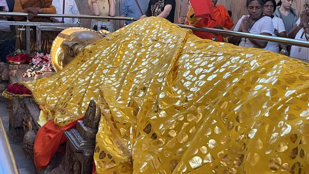 Buddhistische Pilgerreise, Kushinagar. Die Statue einer liegenden Figur, gehüllt in ein goldgemustertes Tuch, umgeben von Menschen in einer Tempelumgebung.
