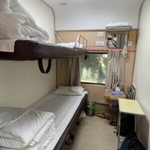 Eine kleine Zugkabine mit zwei übereinander gestapelten Betten, einem Fenster, einem Vorhang, einem kleinen Tisch mit Büchern und Schuhen auf dem Boden.