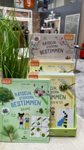 Auf einem Ständer sind Bücher zur Pflanzen- und Insektenbestimmung mit Aufklebern und Puzzles ausgestellt. Im Vordergrund ist eine Pflanze zu sehen.