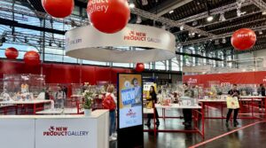 Eine Ausstellungshalle mit Ausstellungen der „New Product Gallery“. Rot-weißes Dekor, in Glasvitrinen ausgestellte Produkte und Besucher, die sich die Stände ansehen. Hängende rote Kugeln mit Text.