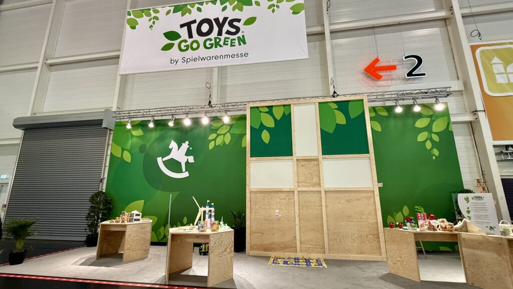 Ausstellungsbereich mit hölzernen Ausstellungstischen, auf denen auf einer Handelsmesse umweltfreundliches Spielzeug unter dem Motto „Toys Go Green“ präsentiert wird.