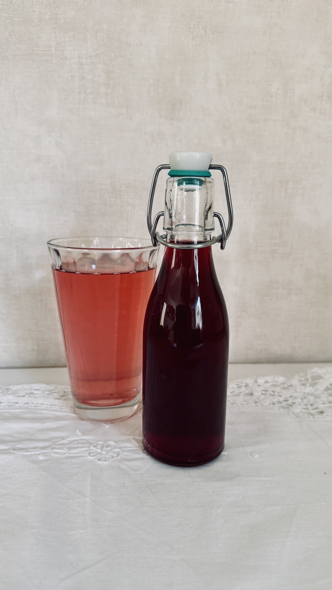 Eine Glasflasche mit dunkelroter Flüssigkeit und verschlossenem Deckel steht neben einem Glas mit einem hellroten Getränk auf einem weißen Tischtuch.