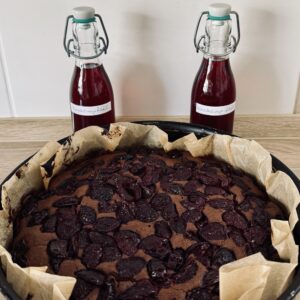 Ein runder, mit Kirschen belegter Schokoladenkuchen steht in Backpapier eingebettet auf einer Holzplatte; hinter dem Kuchen stehen zwei Flaschen mit der Aufschrift "Kirschsirup 2023".
