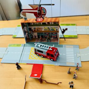 Ein Spielzeug-Feuerwehrset mit einem Feuerwehrauto, einem Hubschrauber, Figuren von Feuerwehrleuten und ausklappbaren Straßen, die auf einem Holztisch angeordnet sind.