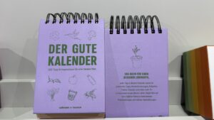 Zwei lilafarbene, spiralgebundene Tischkalender mit dem Titel "Der Gute Kalender", die Illustrationen von Pflanzen, Lebensmitteln und umweltfreundlichen Themen enthalten, stehen in einem Regal.