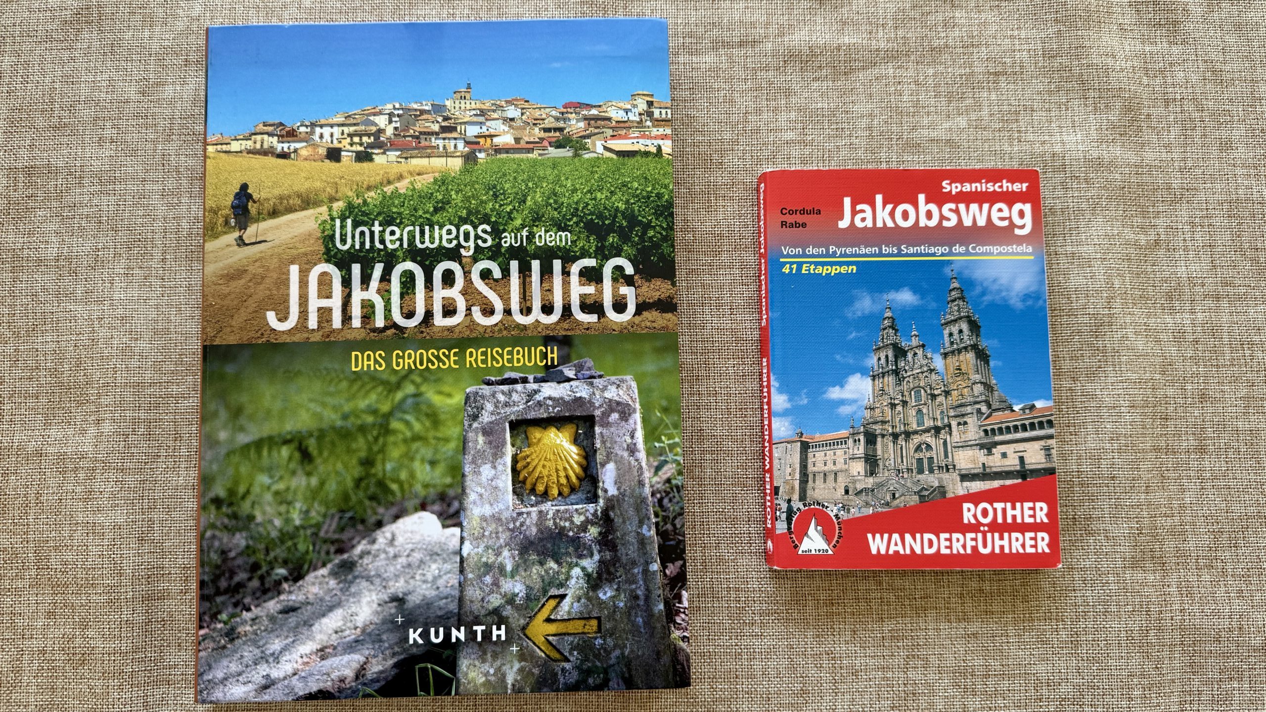 Zwei Reiseführer über den Jakobsweg sind auf einer braunen, strukturierten Oberfläche platziert, einer rot und im Taschenformat, der andere größer mit einem Foto einer Wegmarkierung und eines Dorfes.