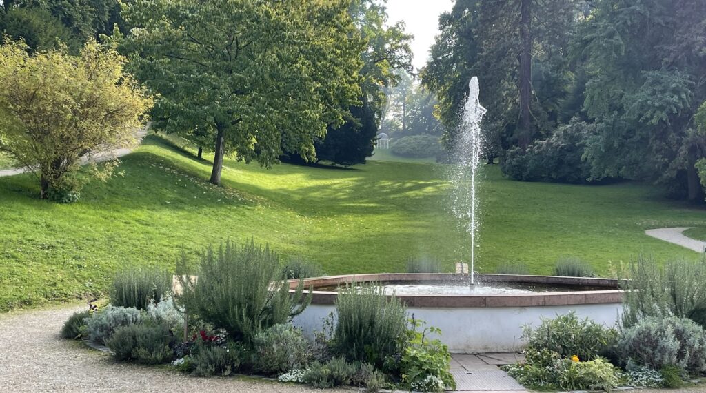 Ein runder Brunnen mit einem zentralen Wasserstrahl ist von Pflanzen und Bäumen in einem üppigen, grünen Park umgeben.
