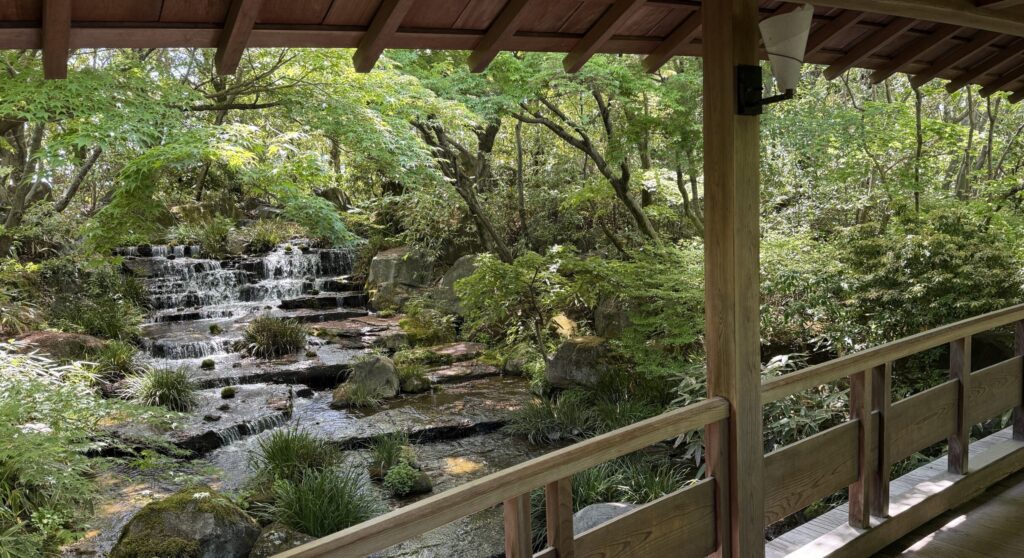 Ein Holzsteg im Koko-En Garten neben Himeji Burg bietet einen Blick auf einen kleinen, rauschenden Wasserfall, der von grünen Bäumen und Felsen inmitten einer ruhigen Gartenlandschaft umgeben ist.