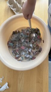 Eine Hand rührt mit einem Löffel in einem Plastikschüssel mit Wasser, in dem zerrissene, bunte Papierstücke einweichen.