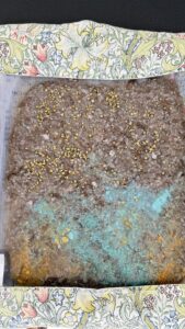 Ein rechteckiges, strukturiertes Material mit braunen, türkisfarbenen und gelben Bereichen, übersät mit kleinen goldfarbenen Partikeln, eingefasst von Stoff mit Blumenmuster.